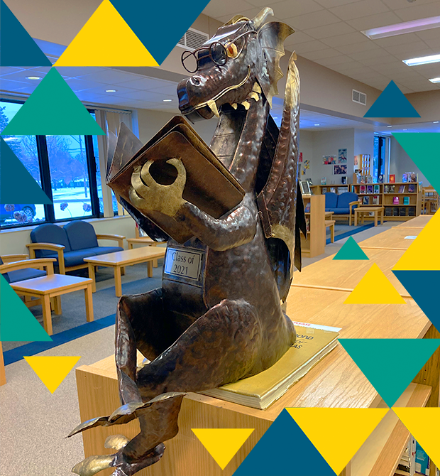 A metal sculpture of a dragon wearing glasses and holding a book sits atop a bookshelf in the Kalkaska High School library. The dragon sculpture bears a plaque that reads "Class of 2021" and there are blue, aqua, and yellow triangles around the edges of the photo.